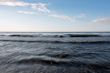 Sakin, mavi bir saatin altında, bulutlar dağılmış, hafif dalgalar. Akşam alacakaranlık deniz manzarası yumuşak, solgun ışığı yansıtan desenli suyla. Sakin ve atmosferik.