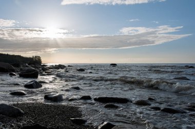 Kıyıya vuran dalgaları olan Rocky plajı parçalı bulutlu bir gökyüzünün altında güneş ışığıyla.
