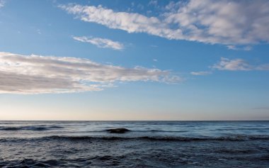 Alacakaranlıkta dağınık bulutlarla mavi bir gökyüzünün altında küçük dalgalarla sakin bir deniz. Bir dalganın koyu gölgesi, yan tarafından dolayı su yüzeyinde görünür ve hafifçe arkadan aydınlatılır..
