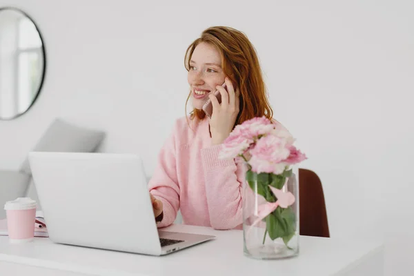 Masada Dizüstü Bilgisayarla Çalışırken Cep Telefonuyla Konuşan Gülümseyen Kızıl Saçlı — Stok fotoğraf