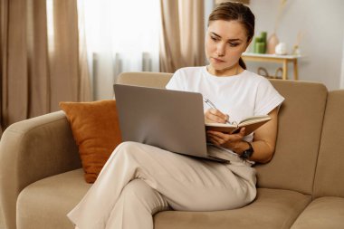 Yetişkin bir kadın evdeki kanepede otururken dizüstü bilgisayar kullanıyor ve günlüğünü yazıyor..