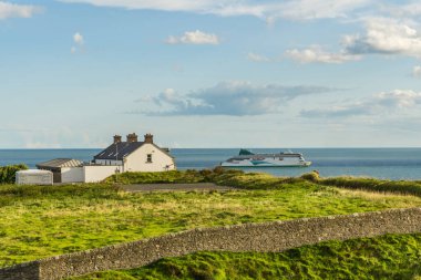 Okyanusa bakan bir dağlık alana tünemiş bir ev ve arka planda bir tekne. Çimenler ve bitkiler bulutlu gökyüzünün altında pitoresk bir doğal manzara oluştururlar.