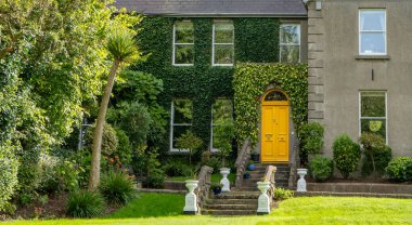 Sarı kapılı büyük bir evin önünde, güzel yemyeşil bir çim var, resim gibi bir manzara yaratıyor.