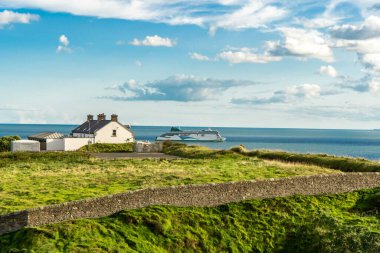 Okyanusa bakan bir dağlık alana tünemiş bir ev ve arka planda bir tekne. Çimenler ve bitkiler bulutlu gökyüzünün altında pitoresk bir doğal manzara oluştururlar.
