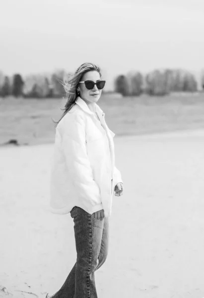 Woman in white jacket, sunglasses and blue jeans at the beach, new collection, fashionable trend