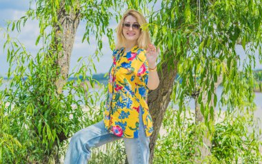 Tatil tarzı, kadın kıyafetleri. Genç Avrupalı bronz tenli, sarı saçlı, kot pantolonlu ve kumsalda renkli pamuklu gömlekli bir kadın.