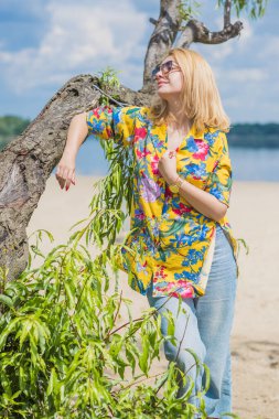 Tatil tarzı, kadın kıyafetleri. Genç Avrupalı bronz tenli, sarı saçlı, kot pantolonlu ve kumsalda renkli pamuklu gömlekli bir kadın.
