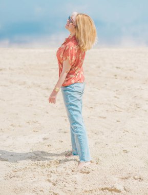Tatil tarzı, kadın kıyafetleri. Genç Avrupalı bronz tenli, sarı saçlı, kot pantolonlu ve kumsalda renkli pamuklu gömlekli bir kadın.