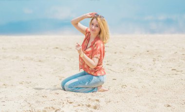 Tatil tarzı, kadın kıyafetleri. Genç Avrupalı bronz tenli, sarı saçlı, kot pantolonlu ve kumsalda renkli pamuklu gömlekli bir kadın.