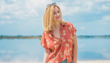 Tatil tarzı, kadın kıyafetleri. Genç Avrupalı bronz tenli, sarı saçlı, kot pantolonlu ve kumsalda renkli pamuklu gömlekli bir kadın.