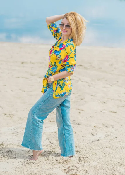 度假风格女装衣橱图片说明在海滩上穿着蓝色牛仔裤和色彩斑斓的棉质衬衫的年轻的欧洲晒黑妇女 — 图库照片