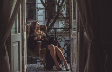 Peignoir giymiş bir kadın ve bir kadeh şarap. Tüylü siyah ipek bornozlu Avrupalı sarışın kadının şehvetli fotoğrafı. Moda konsepti