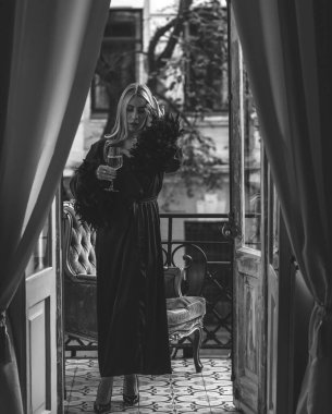 Peignoir giymiş bir kadın ve bir kadeh şarap. Tüylü siyah ipek bornozlu Avrupalı sarışın kadının şehvetli fotoğrafı. Moda konsepti