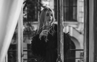 Peignoir giymiş bir kadın ve bir kadeh şarap. Tüylü siyah ipek bornozlu Avrupalı sarışın kadının şehvetli fotoğrafı. Moda konsepti