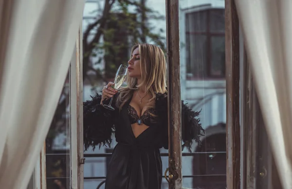 Lady in peignoir with a glass of wine. Sensual photo of European Blonde woman in black silk robe with feathers sleeve. Fashionable concept