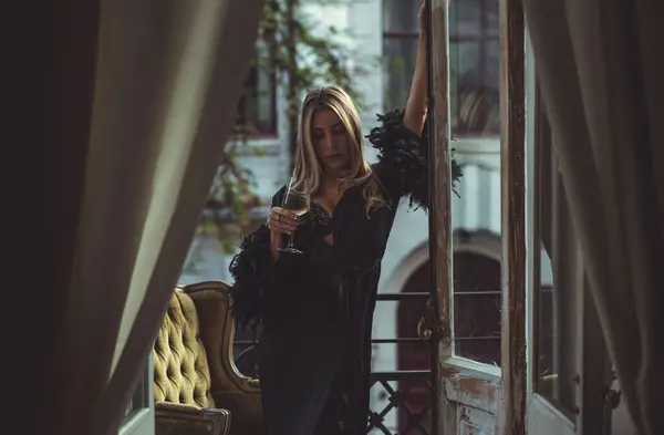 Lady in peignoir with a glass of wine. Sensual photo of European Blonde woman in black silk robe with feathers sleeve. Fashionable concept