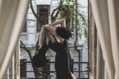 Yatak odasında zarif bir kadın. Siyah ipek bornozlu, balkonda elinde bir bardak şampanyayla duran kollarında tüyleri olan sarışın bir Avrupalı kadın..  