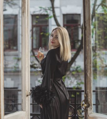 Yatak odasında zarif bir kadın. Siyah ipek bornozlu, balkonda elinde bir bardak şampanyayla duran kollarında tüyleri olan sarışın bir Avrupalı kadın..  