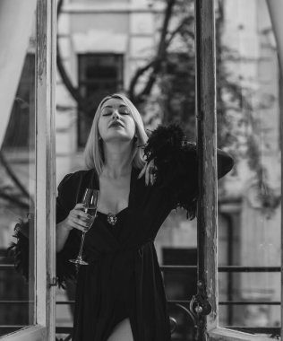 Yatak odasında zarif bir kadın. Siyah ipek bornozlu, balkonda elinde bir bardak şampanyayla duran kollarında tüyleri olan sarışın bir Avrupalı kadın..  