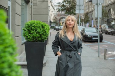 Gri ceketli olgun Avrupalı sarışın kadın şehir merkezinde yürüyor, modaya uygun trendler.