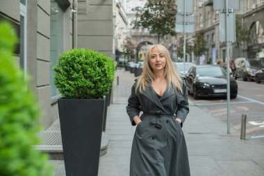 Gri ceketli olgun Avrupalı sarışın kadın şehir merkezinde yürüyor, modaya uygun trendler.