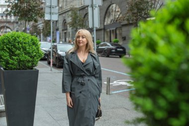 Gri ceketli olgun Avrupalı sarışın kadın şehir merkezinde yürüyor, modaya uygun trendler.