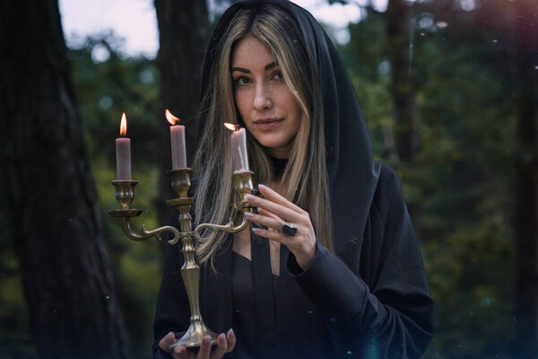 Элегантная серая ведьма в черном плаще держит свечи в мистическом лесу
