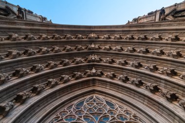Gotik tarzda Katolik bir katedralin girişinin kemerli kemerleriyle meleklerin, azizlerin, yazıtların siluetlerinin bulunduğu açık mavi gökyüzünün arka planına karşı. Vitraylı camla
