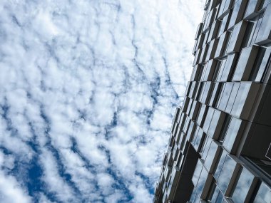 Pencerelerinde bulut yansımaları olan bir binanın köşegen manzarası modern mimari ve kentsel gelişimi parlak, geometrik tarzda sergiliyor. Yatay
