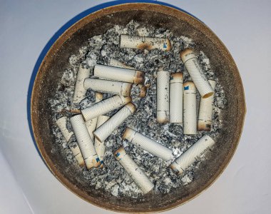 Kötü bağımlılık. Kül tablası ve sigaralar yakın plan. Masadaki kül tablasının yakın görüntüsü. Bir sürü yanmış sigara izmariti ve biraz da kül. Kül tablasında yanan sigaranın tepesinde.