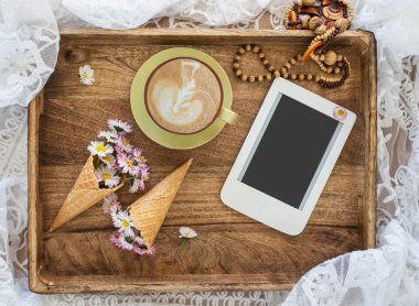 Composition with waffle cone with  flower bouquet, cup of coffee and e-reader on wooden background. Template for feminine blog social media.