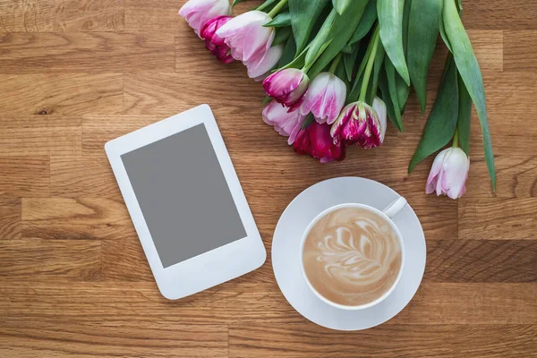 Cup of coffee with pink tulips and e-reader on wooden background.