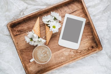 Bir fincan kahveli romantik kompozisyon, çiçek buketli waffle konileri ve tahta tepside fotokopi alanı olan e-okuyucu. Kadınsı blog sosyal medya şablonu.