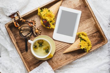 Bir fincan sağlıklı bitki çayı, waffle külahında sarı karahindiba ve ahşap tepside e-kitap. Bitkisel ilaçlar. Düz stil. Kadınsı blog sosyal medya şablonu. 