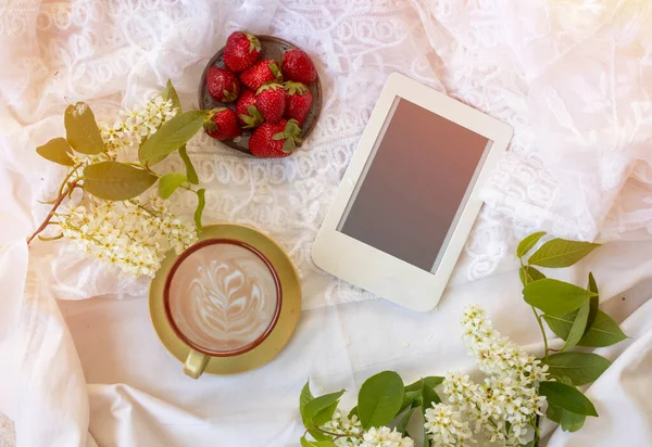 Top view of coffee in bed with tasty strawberries, e-reader and bird cherry branches.