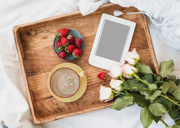 Top view of coffee in bed with tasty strawberries, e-reader and bouquet of roses on wooden tray. Spring, summer still life composition. Romantic surprise.