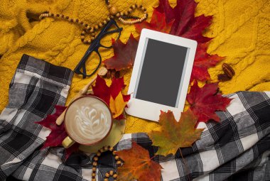 Autumn home cozy composition with checkered plaid, warm sweater, cup of coffee, fall leaves and e-book with copy space. Fall season template for feminine blog social media.