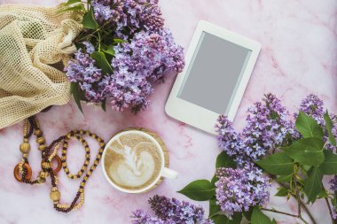 Bir fincan kahveyle rahat kompozisyon, leylak çiçekleri ve pembe mermer arka planda fotokopi alanı olan e-okuyucu. Kadınsı blog sosyal medya şablonu.
