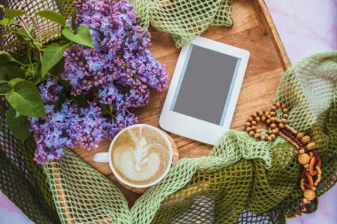 Bir fincan kahveyle rahat kompozisyon, leylak çiçekleri ve ahşap tepside fotokopi alanı olan e-okuyucu. Kadınsı blog sosyal medya şablonu.