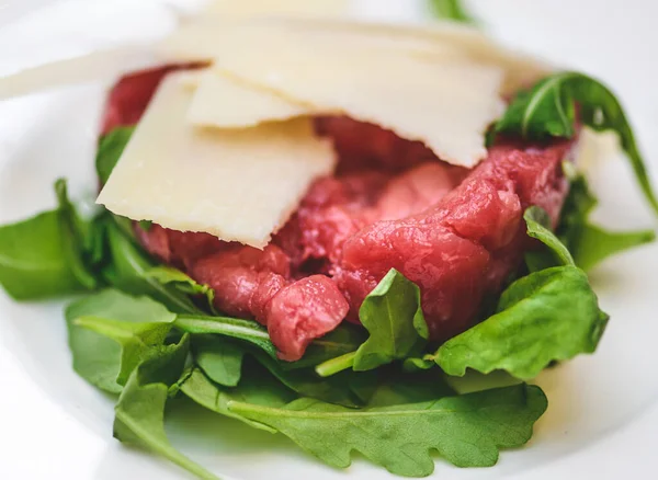 Beef tartare or tartar steak, dish of raw ground minced beef with parmesan cheese flakes and rocket leaves just served in a white plate, close up
