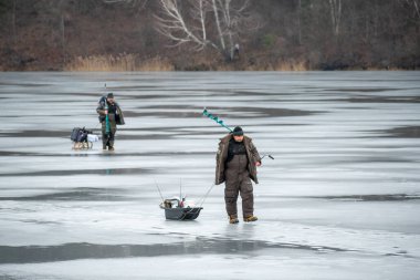 Vilnius, Lithuania - January 15 2023: Fishermen leaving after fishing on a frozen lake in winter with fishing pole or rod, ice auger and equipment for fishing, portable thermal tent on background