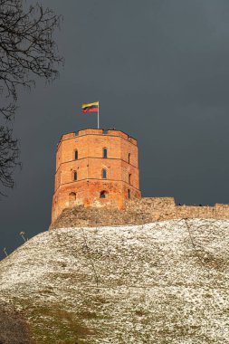 Gediminas Tower or Castle, the remaining part of the Upper Medieval Castle in Vilnius, Lithuania with Lithuanian flag in winter day with snow and beautiful dramatic grey cloudy sky, vertical
