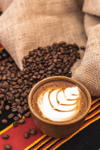 Cappuccino in a ceramic cup with flower or leaves drawn on the foam and roasted coffee beans or seeds scattered on a tablecloth with African decorations and in a jute sack, vertical