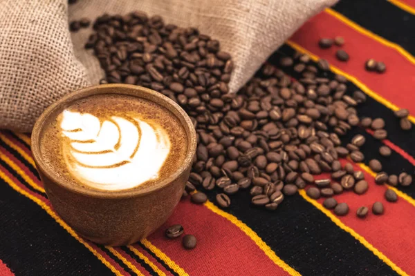 Cappuccino in a ceramic cup with flower or leaves drawn on the foam and roasted coffee beans or seeds scattered on a tablecloth with African decorations and in a jute sack