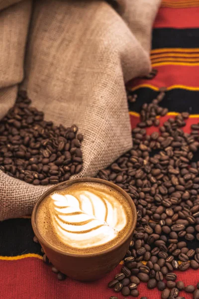 Cappuccino in a ceramic cup with flower or leaves drawn on the foam and roasted coffee beans or seeds scattered on a tablecloth with African decorations and in a jute sack, vertical