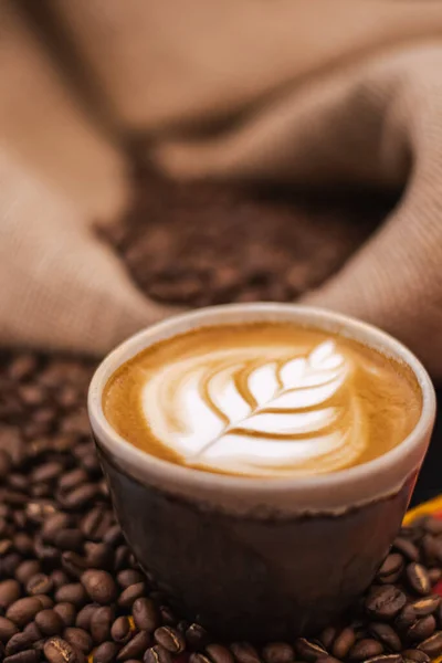 Cappuccino in a ceramic cup with flower or plant drawn on the foam and roasted coffee beans or seeds scattered on the table and in a jute sack, vertical