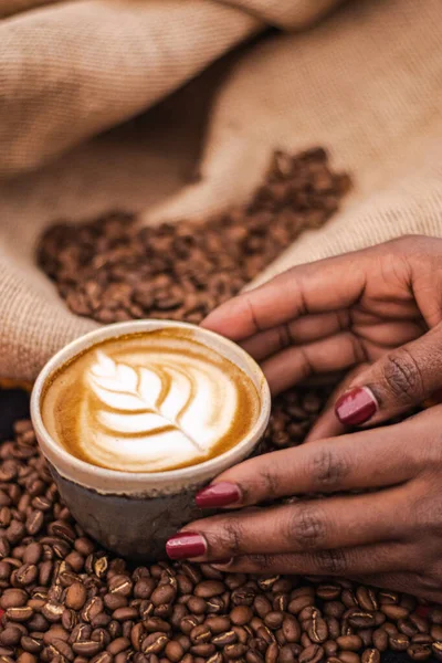 Black skin girl holding a Cappuccino in a ceramic cup with flower or leaves drawn on the foam, teapot and roasted coffee beans or seeds in a jute sack, vertical