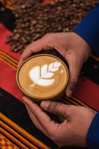 Black skin hand preparing a Cappuccino in a ceramic cup with flower or leaves drawn on the foam and roasted coffee beans or seeds scattered on a tablecloth with African decorations, vertical