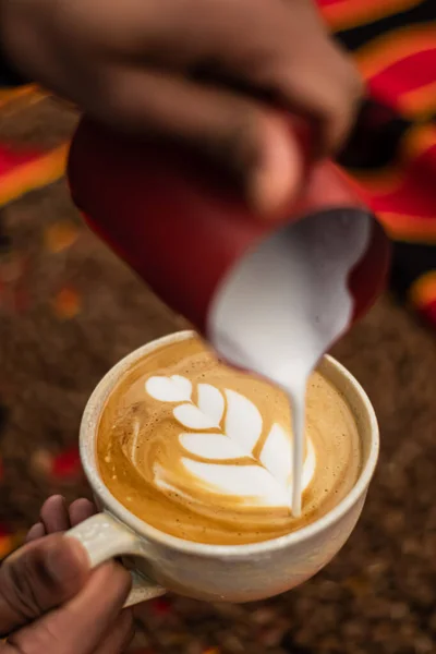 Black skin hand preparing a Cappuccino in a ceramic cup with flower or leaves drawn on the foam and roasted coffee beans or seeds scattered on a tablecloth with African decorations, vertical