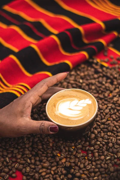 Black skin girl holding a Cappuccino in a ceramic cup with flower or leaves drawn on the foam in the hand of a black skin girl and roasted coffee beans or seeds scattered on a tablecloth, vertical